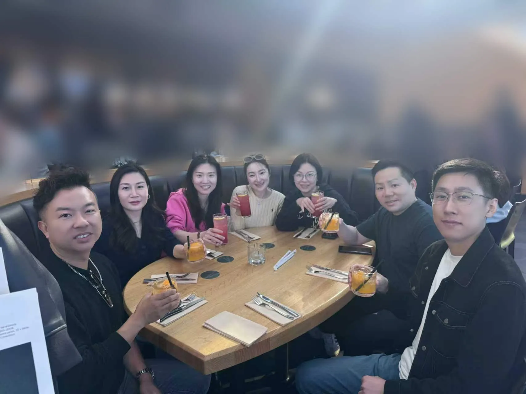 Seven team members raise drinks around a restaurant table; unrelated people behind them are blurred